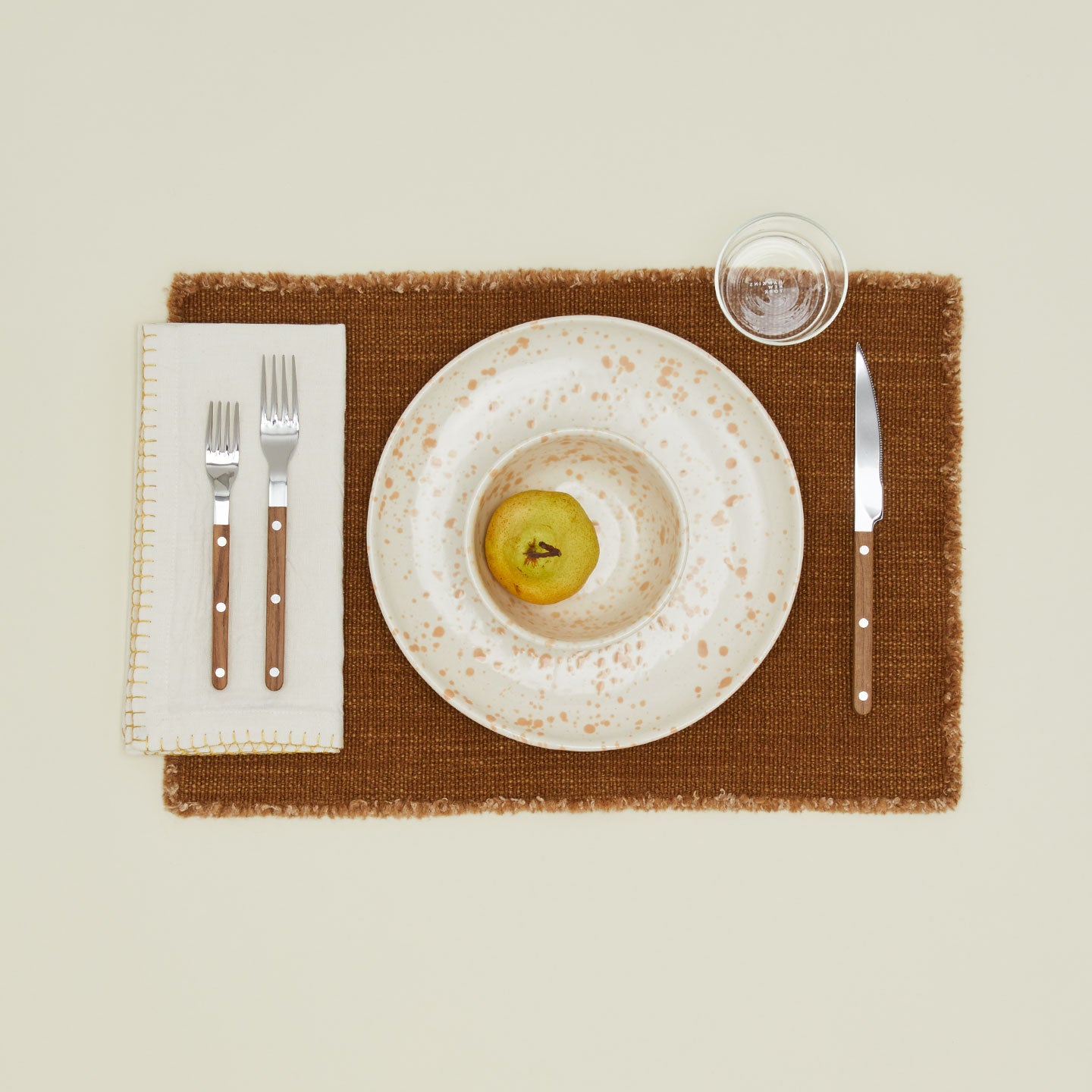 A place setting with a blanket stitched napkin, teak flatware, a splatterware plate and bowl, and a bronze wool placemat. 