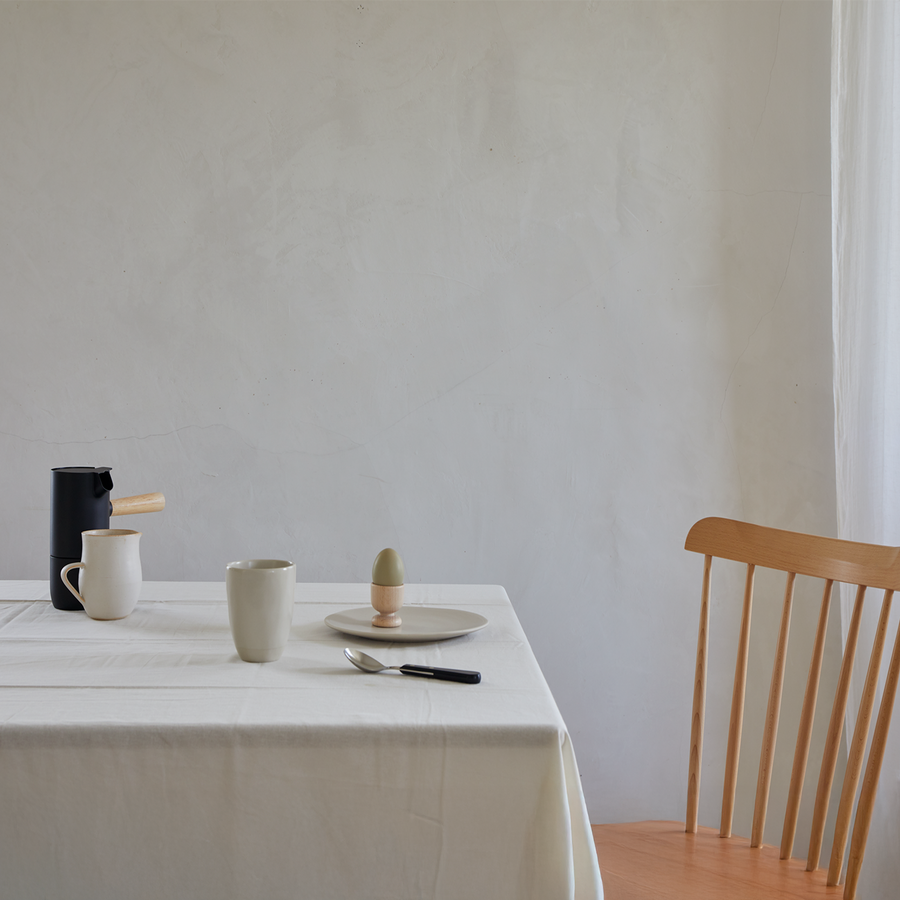 A minimal dining scene with a white tablecloth, a wooden chair, a black coffee pot, a white mug, a plate with an egg in an egg cup, and a knife and spoon on the table.