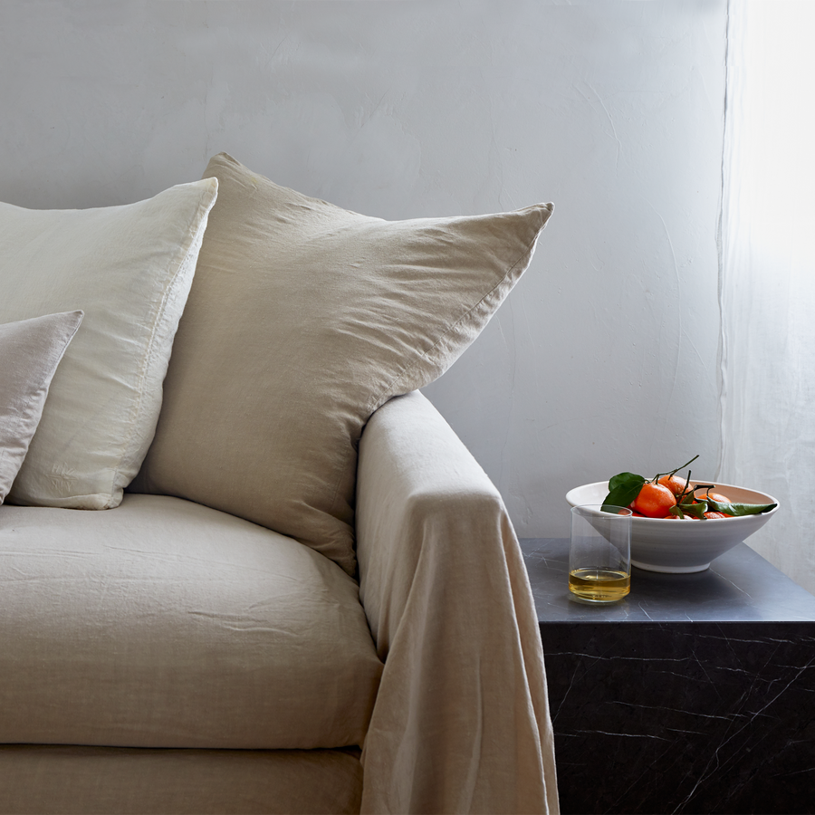 Sofa draped in flax colored linen with neutral linen pillows arranged along the back. Nearby a black marble side table with a bowl of oranges and a tumbler of wine.