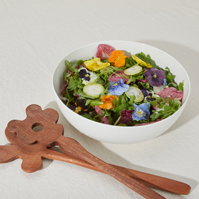 A bowl of salad greens garnished with colorful edible flowers. Nearby sits a pair of carved wood salad servers in floral shapes.