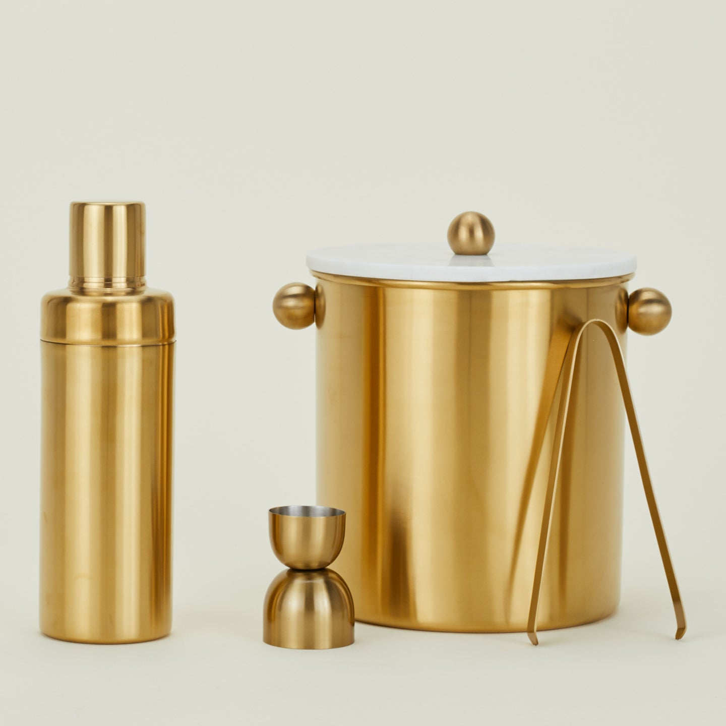 Gold-colored PVD coated stainless steel cocktail shaker, jigger, tongs and ice bucket with white marble lid.